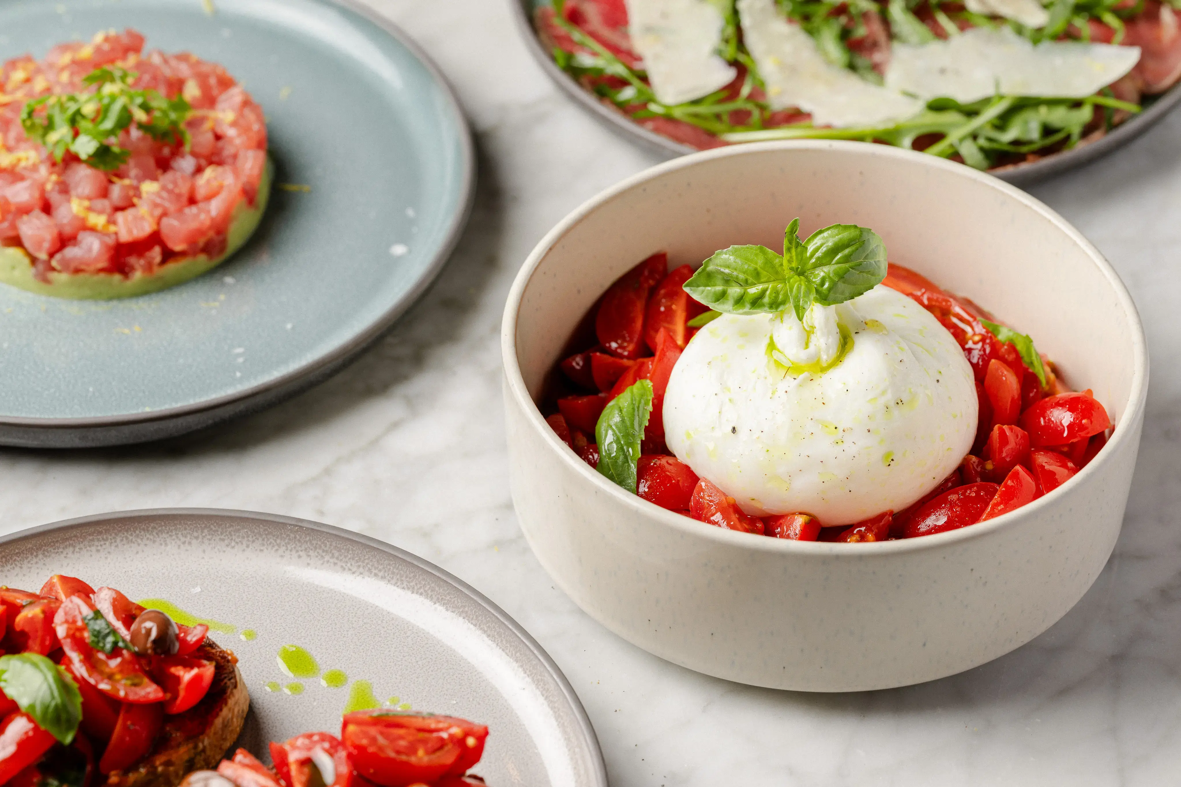 Selection of Italian starters at O Ver Borough, including a burrata caprese salad with fresh basil and Datterino tomatoes, served alongside tuna tartare and bruschetta on a marble table.