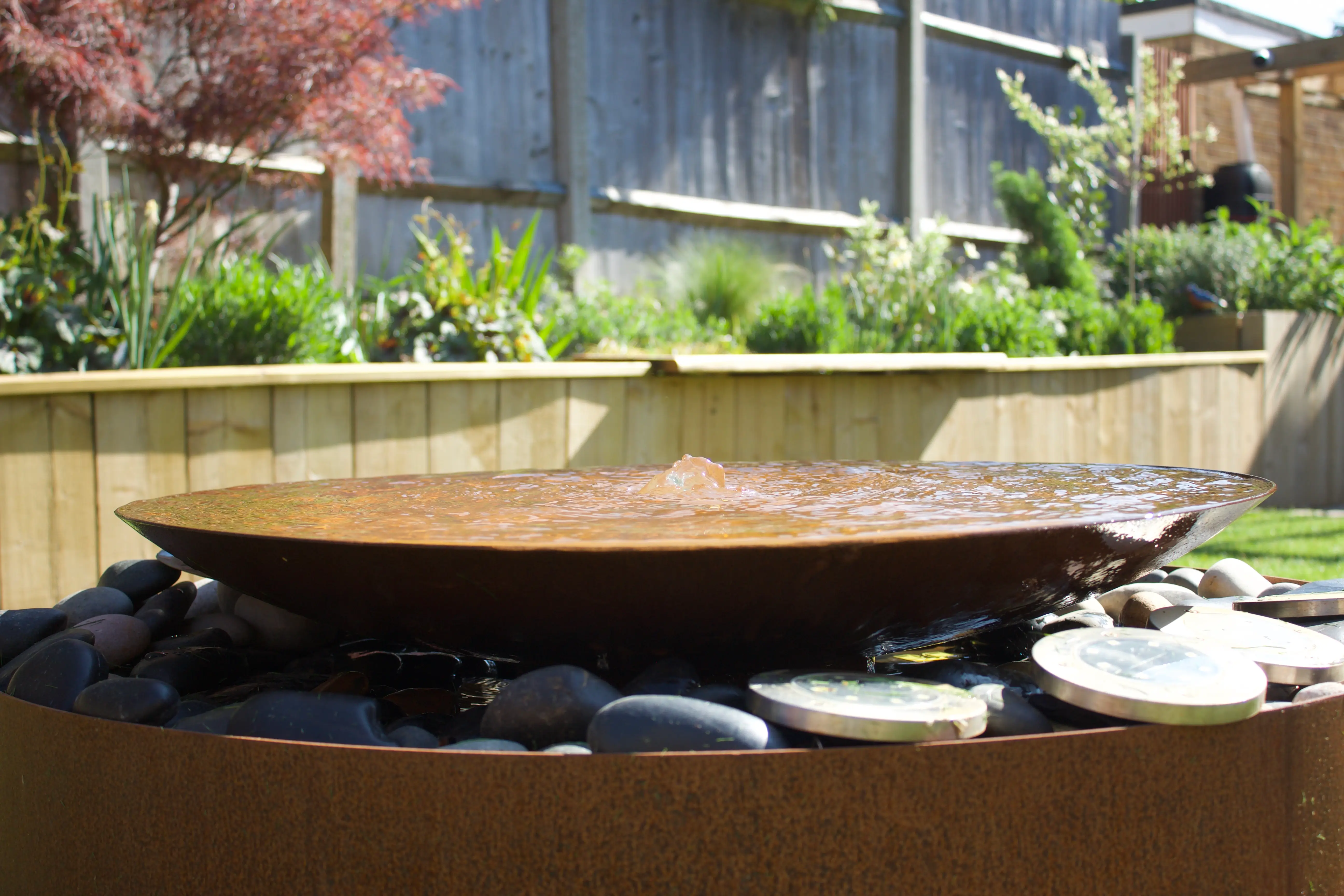 A wooden hot tub surrounded by greenery and a stone border in a serene outdoor setting.