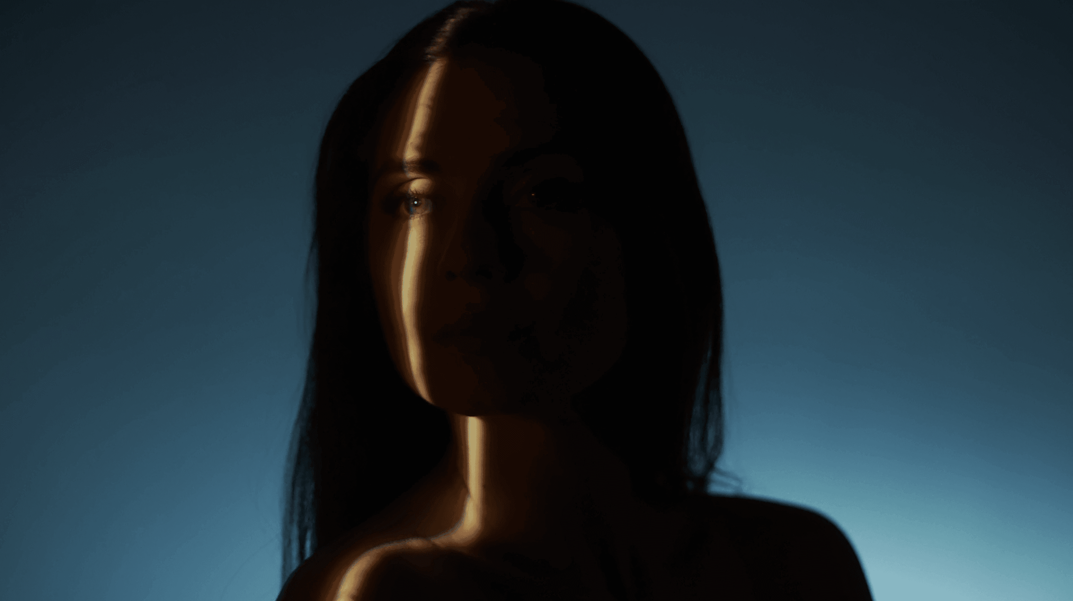 In a softly lit, atmospheric image, a silhouette of a person with long hair stands against a gradient blue background, evoking a sense of mystery and introspection.