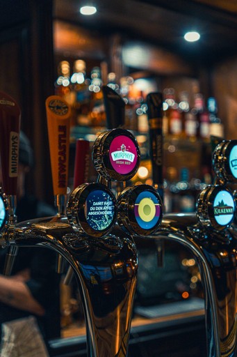 A bottle of beer next to a pint glass on a bar, with green lighting and blurred shelves of bottles in the background.