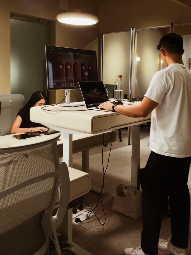 A modern coworking space with a standing desk and a seated workstation, featuring a man standing and working on a laptop while a woman is seated at a nearby desk, both engaged in their tasks.