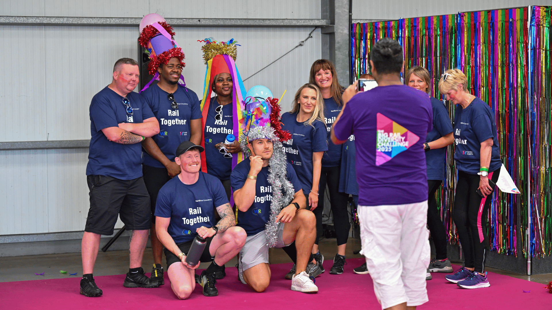 Outdoor team-building event with participants wearing matching blue shirts, taking part in inflatable obstacle courses and games, smiling and cheering, with the words 'ENTER A TEAM' in large bold white and green text across the image.