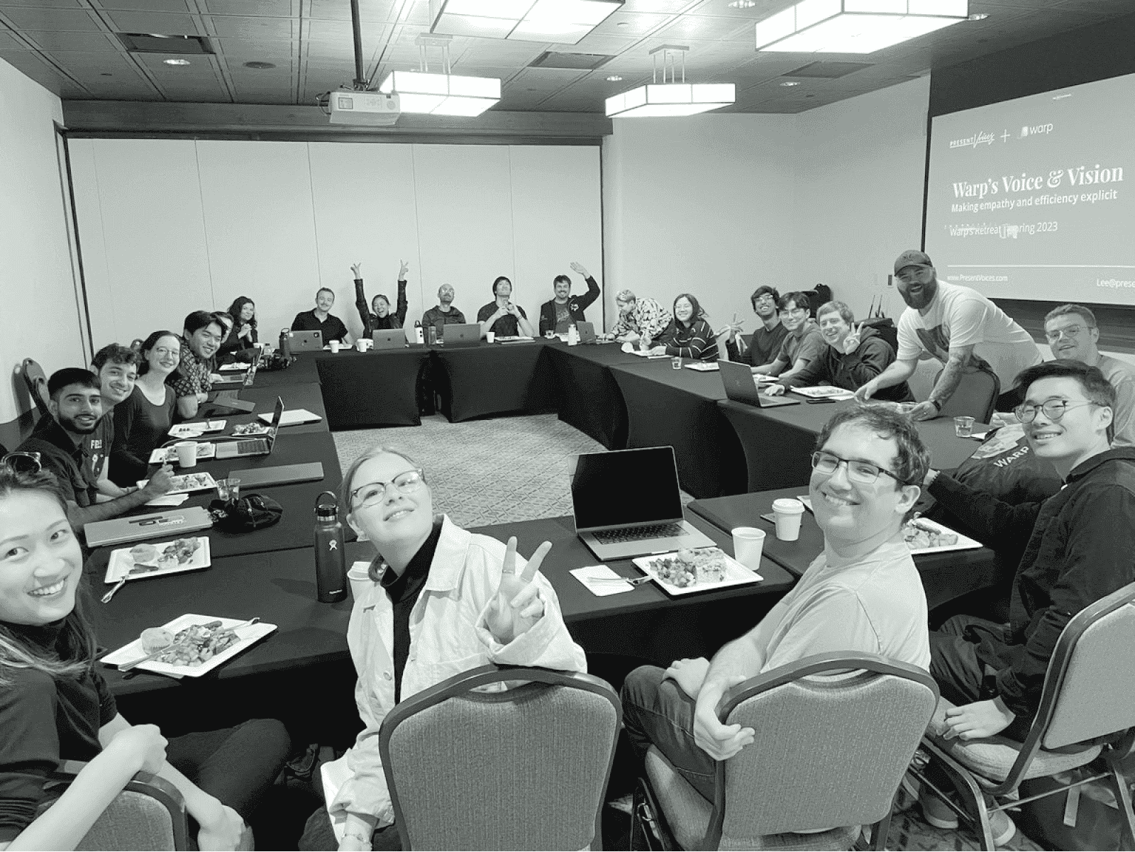 Warp team retreat in a conference room with a presentation screen showing "Warp's Voice & Vision" and attendees smiling at the camera.