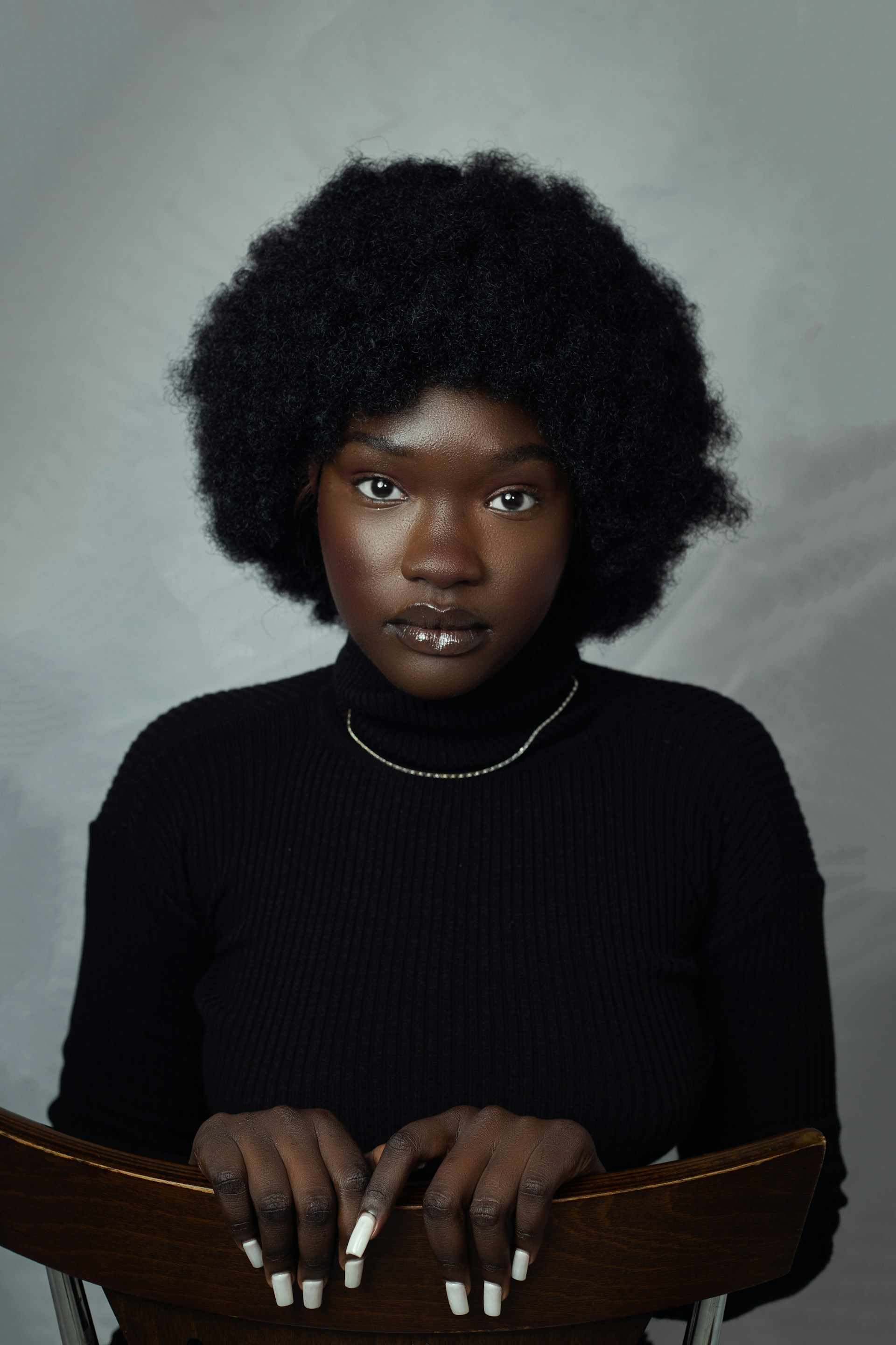 A portrait of a Black woman with a rounded afro, wearing a black turtleneck sweater and a delicate silver necklace, resting her hands on the back of a wooden chair.