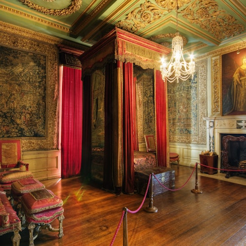 Koristeellinen huone, jossa on kattokruunu, pylvässänky punaisilla verhoilla, antiikkihuonekaluja, kuvioituja seinävaatteita ja takka.