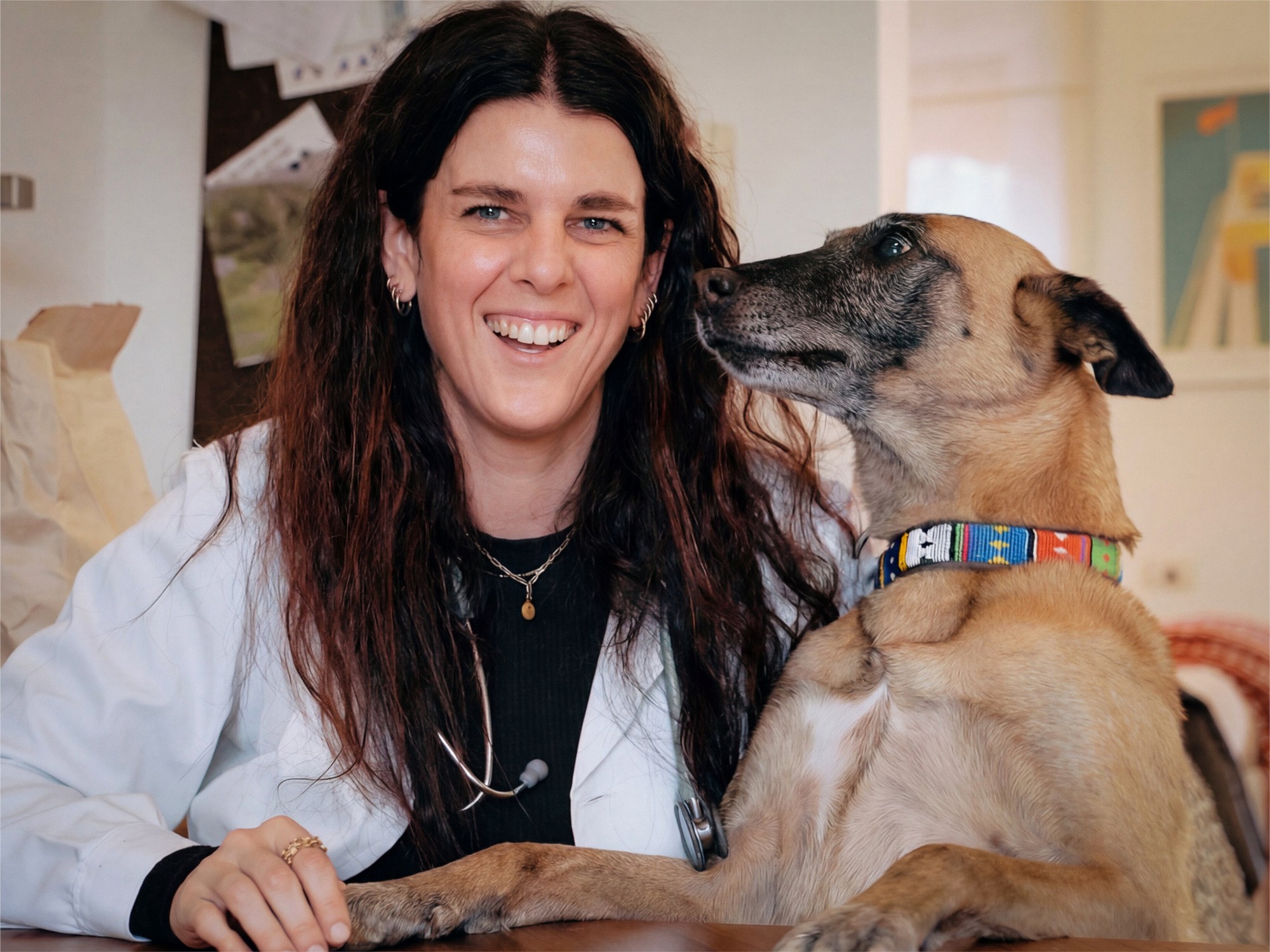 A smiling veterinarian sitting with a friendly dog, both looking at the camera in a cozy clinic setting.