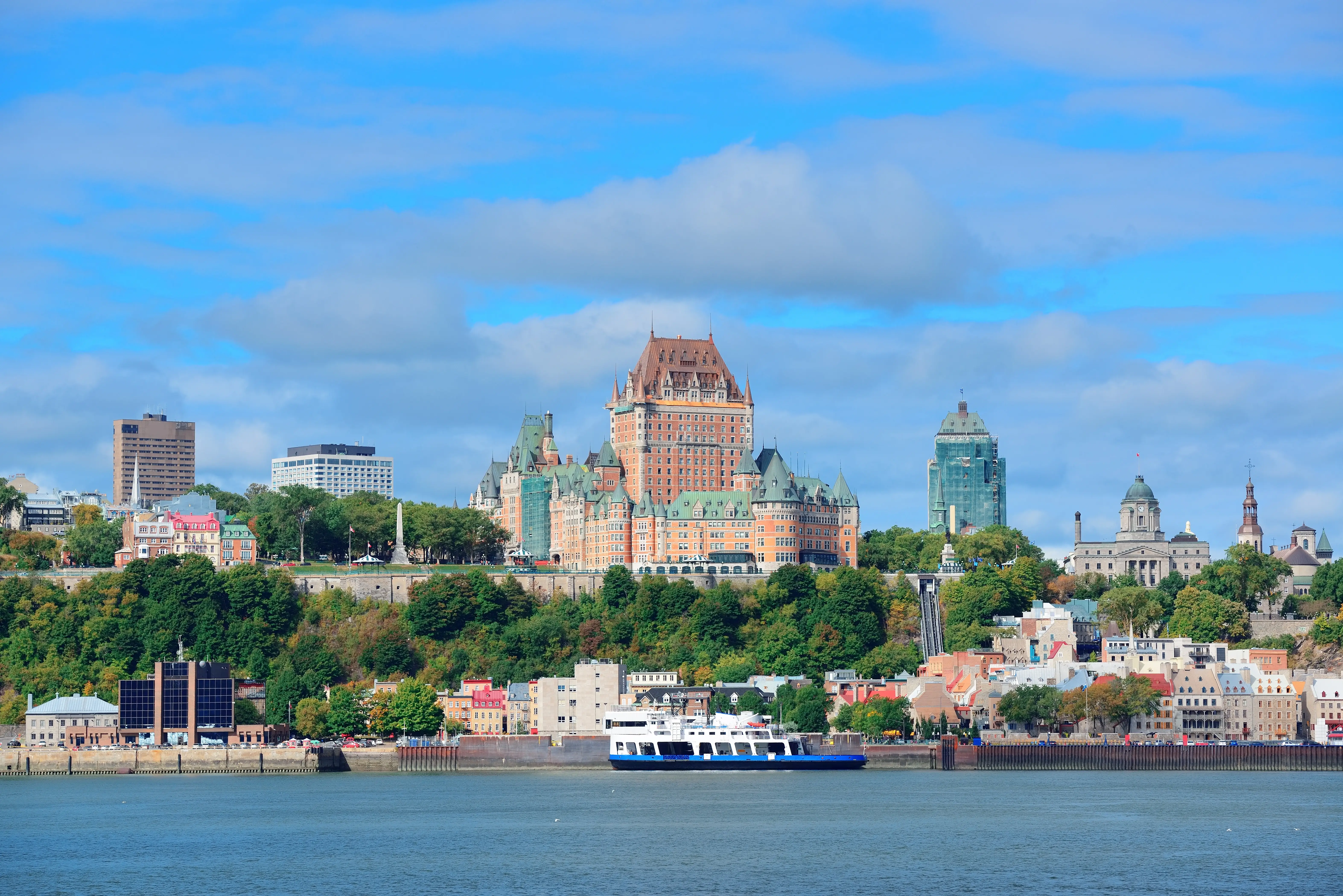 Quebec City skyline with Château Frontenac overlooking the St. Lawrence River, featuring historic buildings, greenery, and a boat on the water under a blue sky.