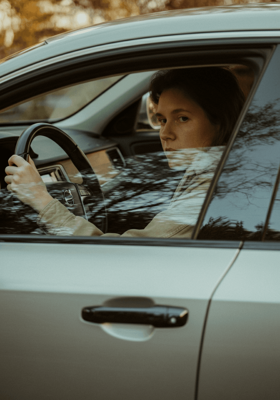 Woman driving a car. She's looking into the camera with a serious expression, holding the steering wheel.