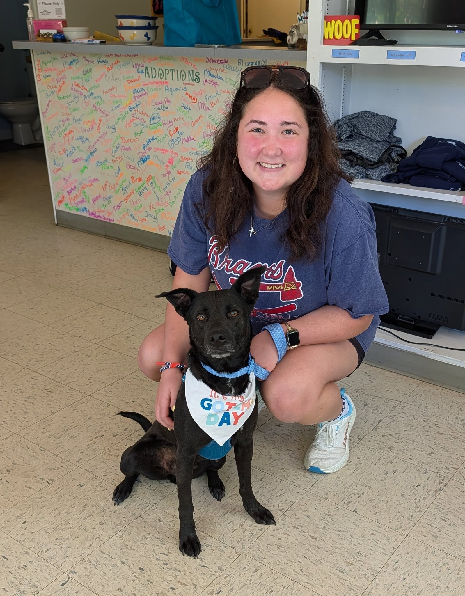 A person sits on the floor beside a small black dog wearing a harness, smiling in a bright indoor space.