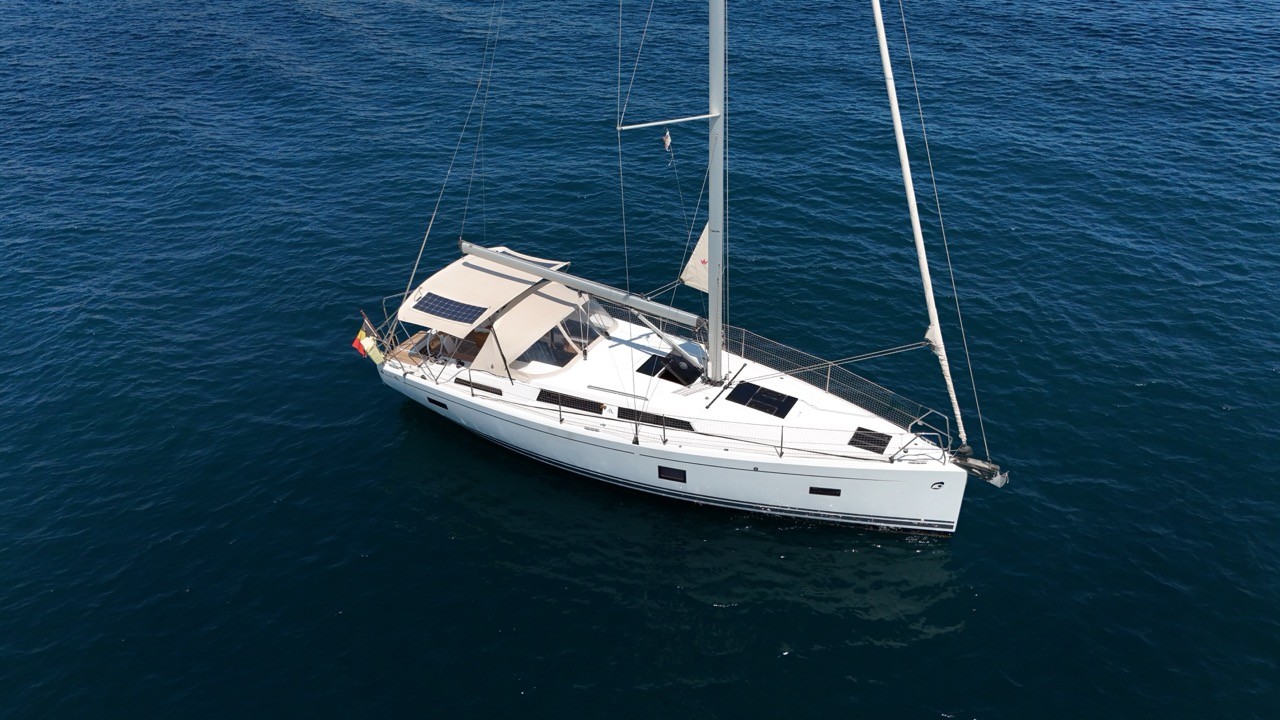 Aerial side view of a Hanse 388 sailing yacht at anchor, showing its self-tacking jib track, solar panels mounted on the bimini, and clean foredeck against dark blue water.
