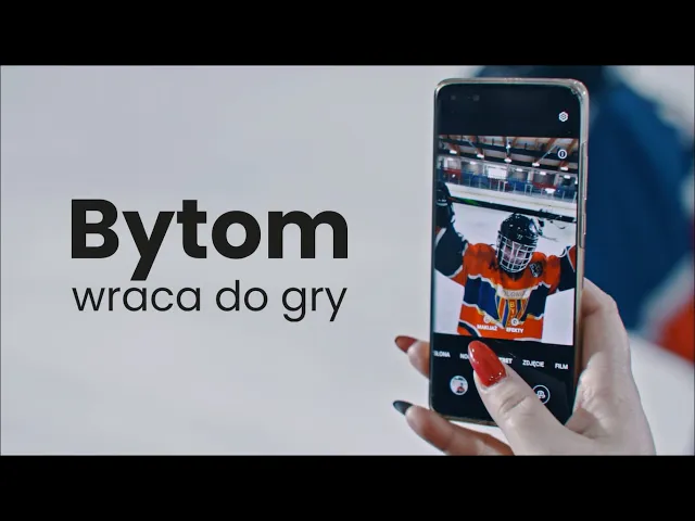 Budynek z wieloma oknami, prawdopodobnie szkoła lub biuro. Zielony pas na dole budynku i kamienny murek.