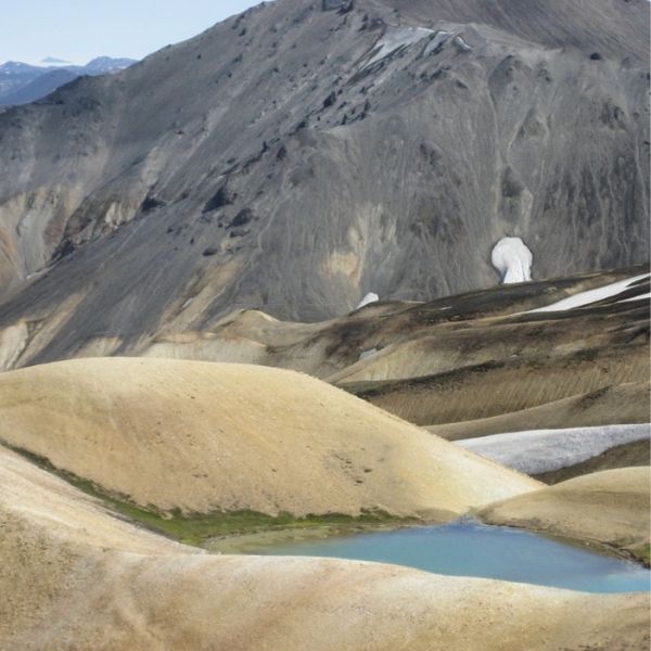 A hiker’s silhouette in the foreground of an aerial view of Landmannalaugar, which is at the north end of the Torfajökull volcanic system. Winding rivers snake through the landscape and moss- and snow-covered red mountains are in the background. 