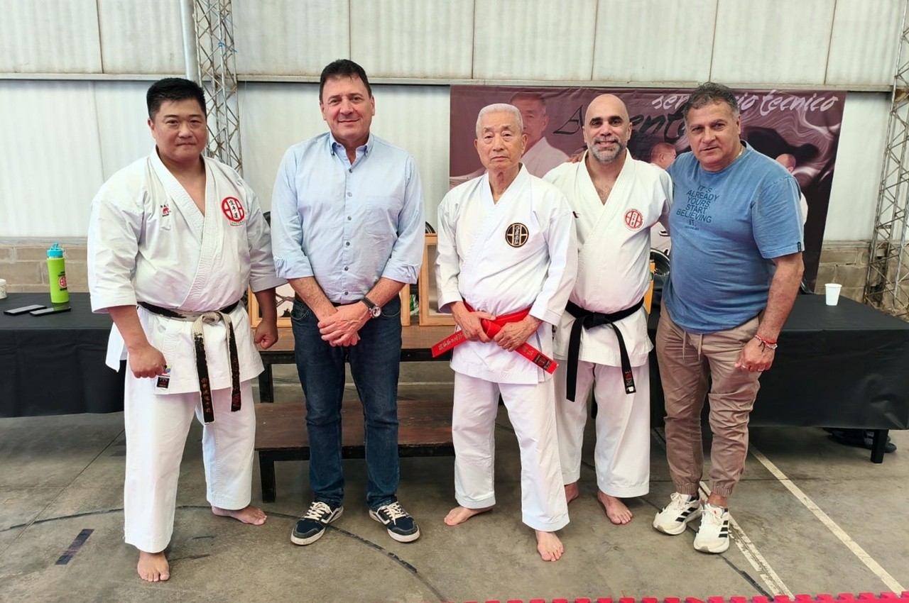 Cinco hombres posan en un seminario de karate, con banner conmemorativo al fondo.