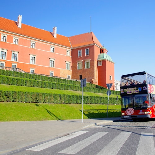 A double-decker tour bus passes by a historical red building with green hedges in front on a sunny day.