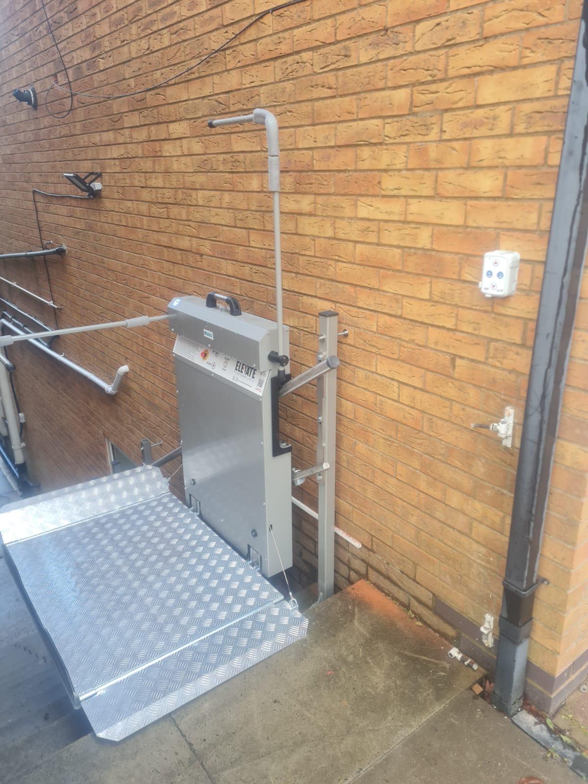Elevate inclined platform stairlift installed at ground level against a brick wall — aluminium chequerplate platform in lowered position with wall-mounted control unit, outdoor commercial installation