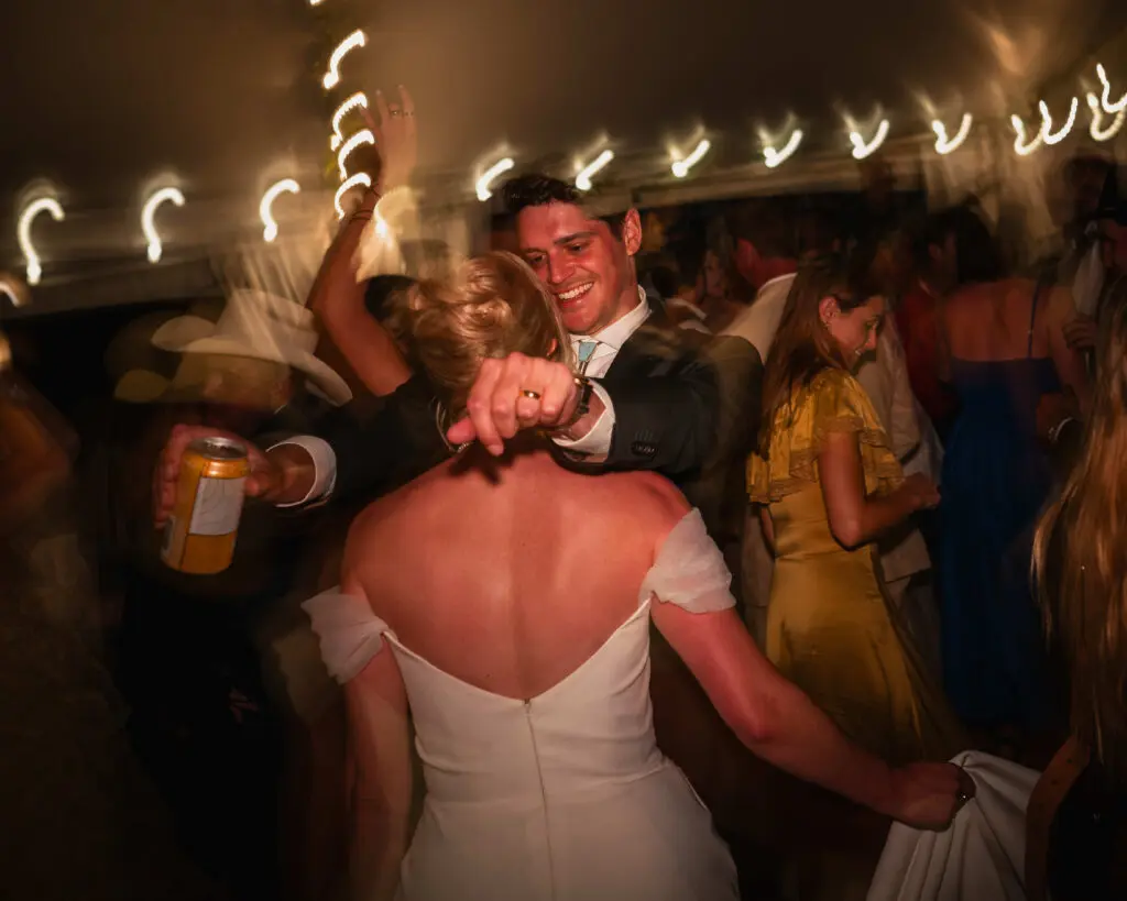 Newlyweds dancing energetically at evening reception with motion blur light trails and guests surrounding couple.