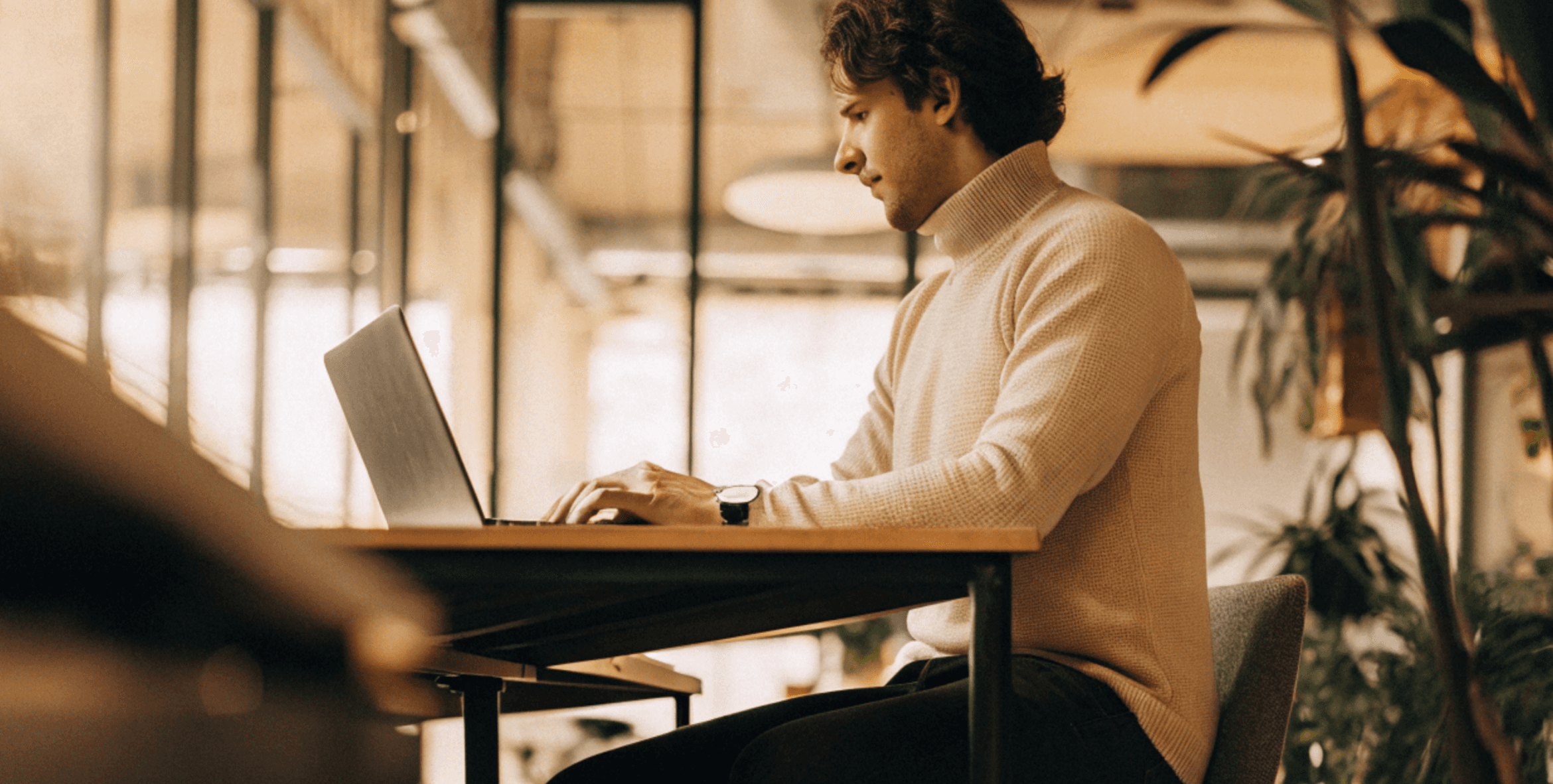 Man in a turtleneck sweater working on a laptop at a desk, side view, indoors with plants and windows.