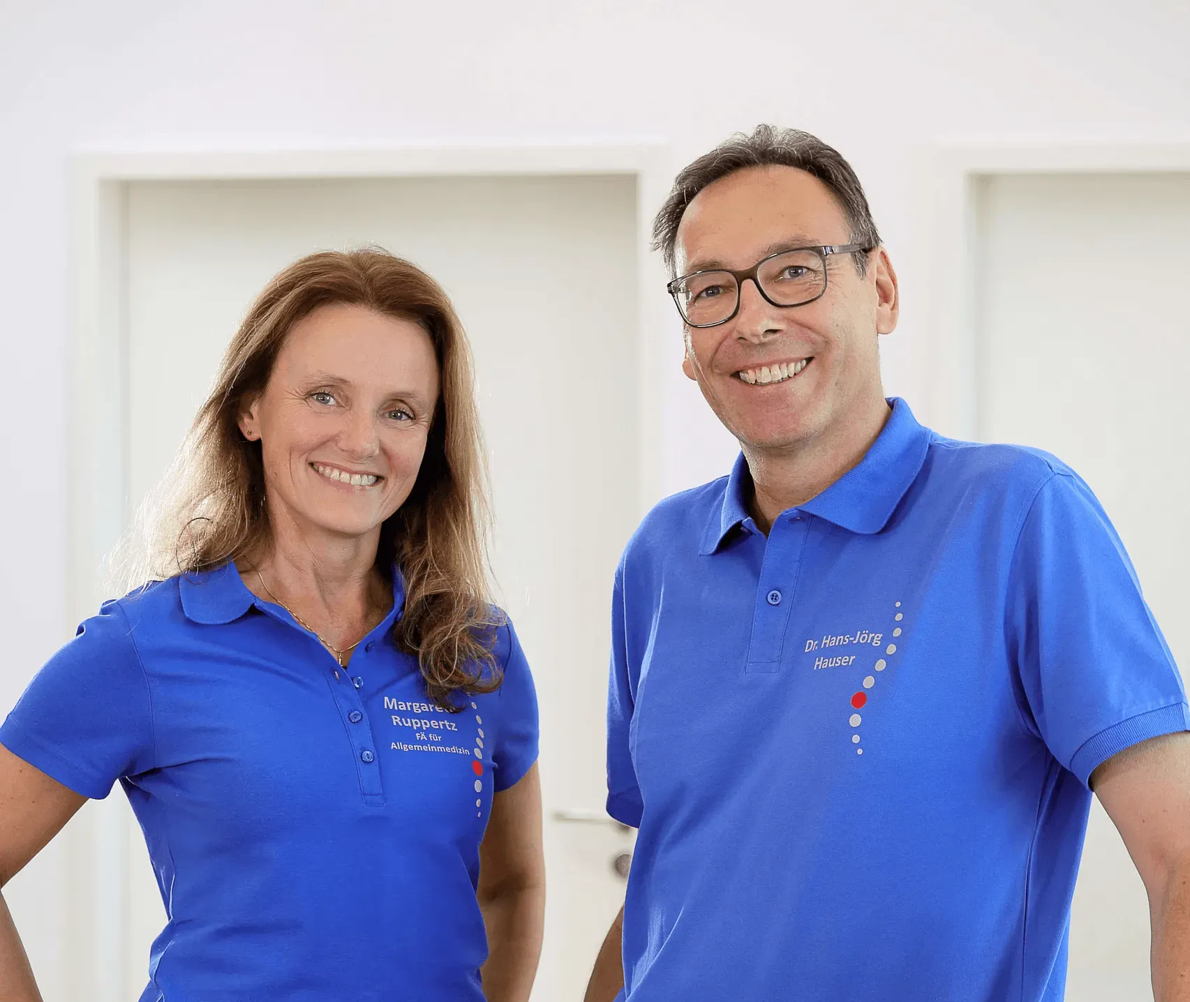 Two smiling individuals in blue shirts stand together in a bright, neutral-colored room.