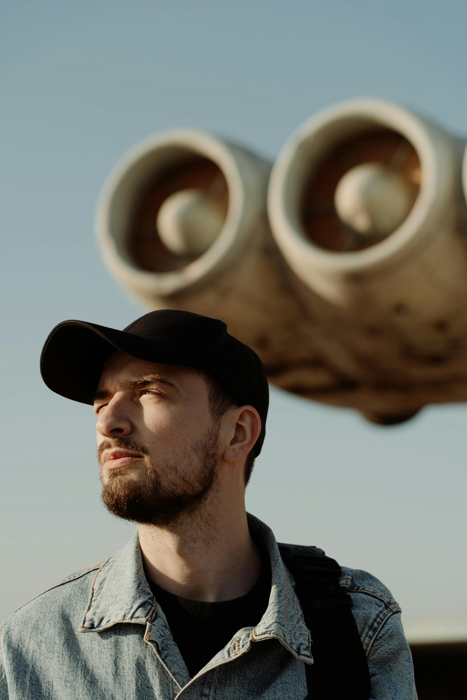 A close-up shot of a man with a beard and a black baseball cap, looking off to the side with large, out-of-focus jet engines in the background.