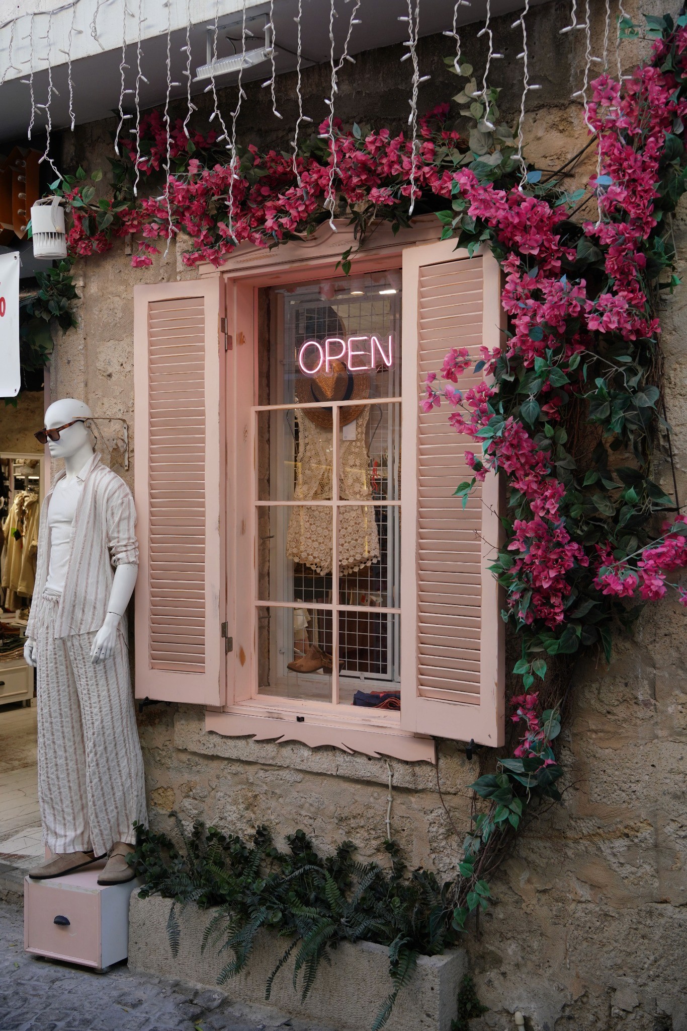 A pink-shuttered boutique window framed by bright bougainvillea glows with an OPEN sign beside a mannequin.