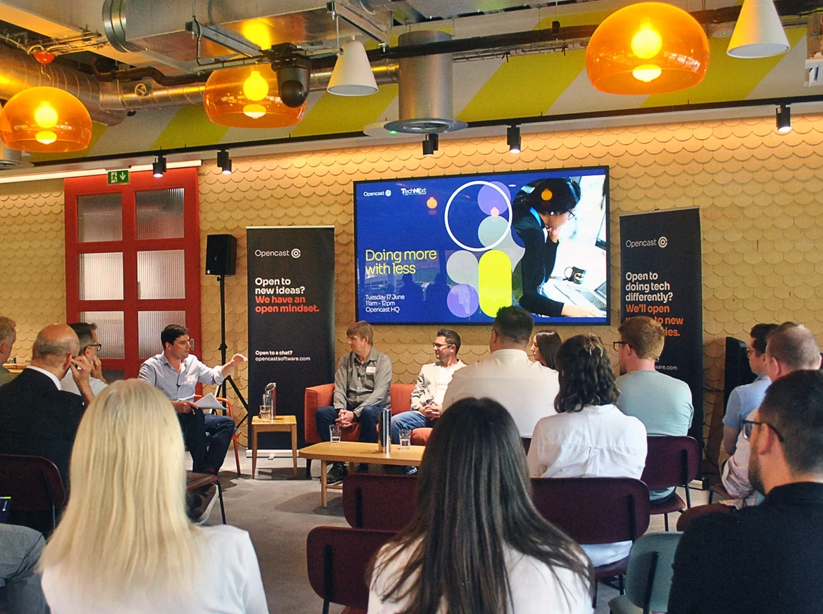 Audience seated facing a panel discussion in a modern event space, with banners and a large screen displaying “Doing more with less” and colourful graphics.