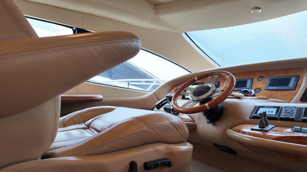 Azimut 68 Evolution lower helm station showing the branded wood-rim stainless steel steering wheel, burl walnut instrument console with Simrad chartplotters and engine gauges, joystick docking control, and tan leather captain seat with panoramic windscreen forward