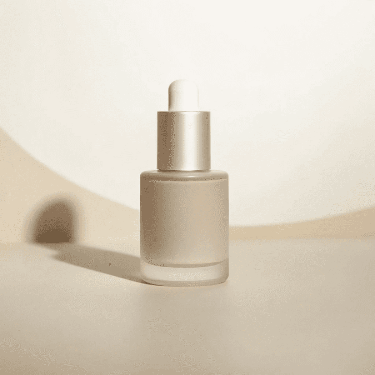 A minimalist glass dropper bottle filled with a light cream-colored serum is set against a softly lit, neutral beige background, casting a subtle shadow; perfect for a skincare, beauty, or cosmetic product context.
