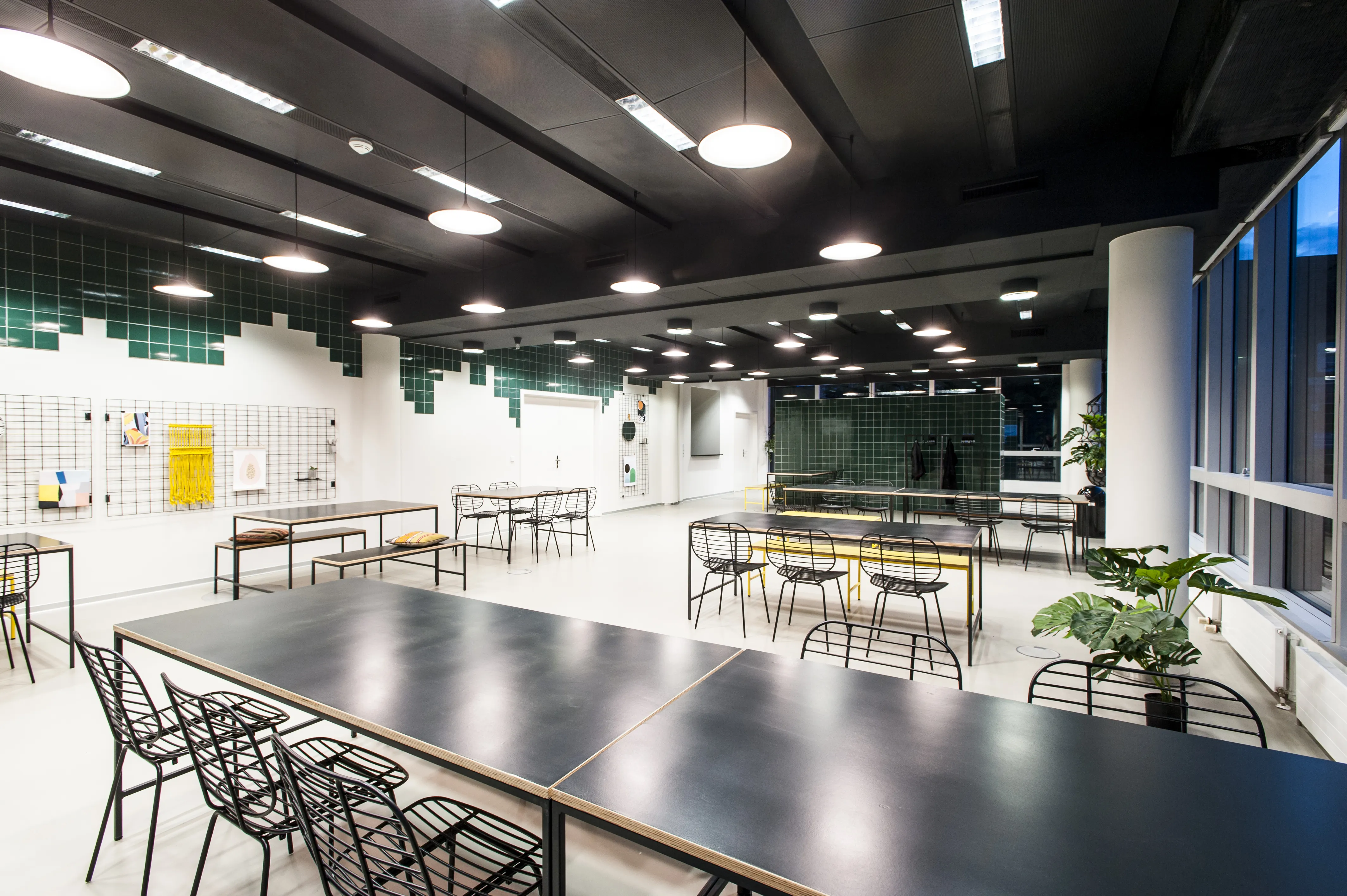 Overview of the KANTINE KOMTUR canteen with black wire chairs and dark tables, green pixel-pattern tile walls, wire grid panels and round pendant lights on a black ceiling.