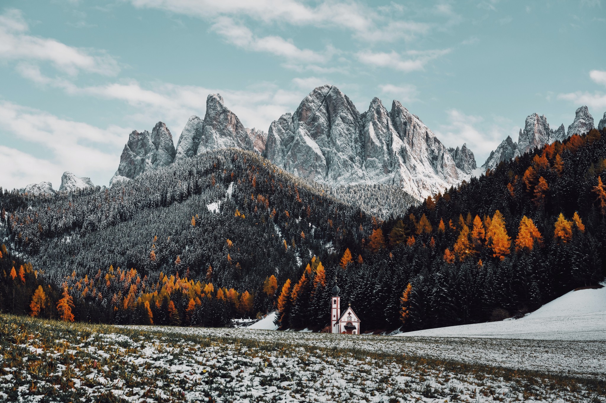 A chapel in a snowy field with black and orange trees and mountains in the background