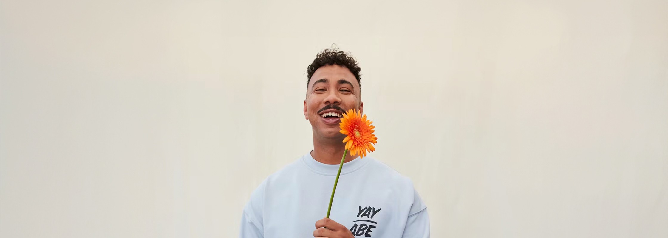 A medium shot of Yay Abe holding an orange flower next to his face, smiling fromn cheek to cheek