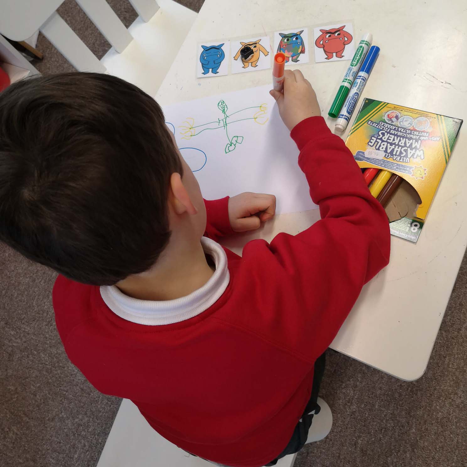 A child sits at a desk and is colouring and drawing on paper, they're using larger colouring pens to draw monsters.