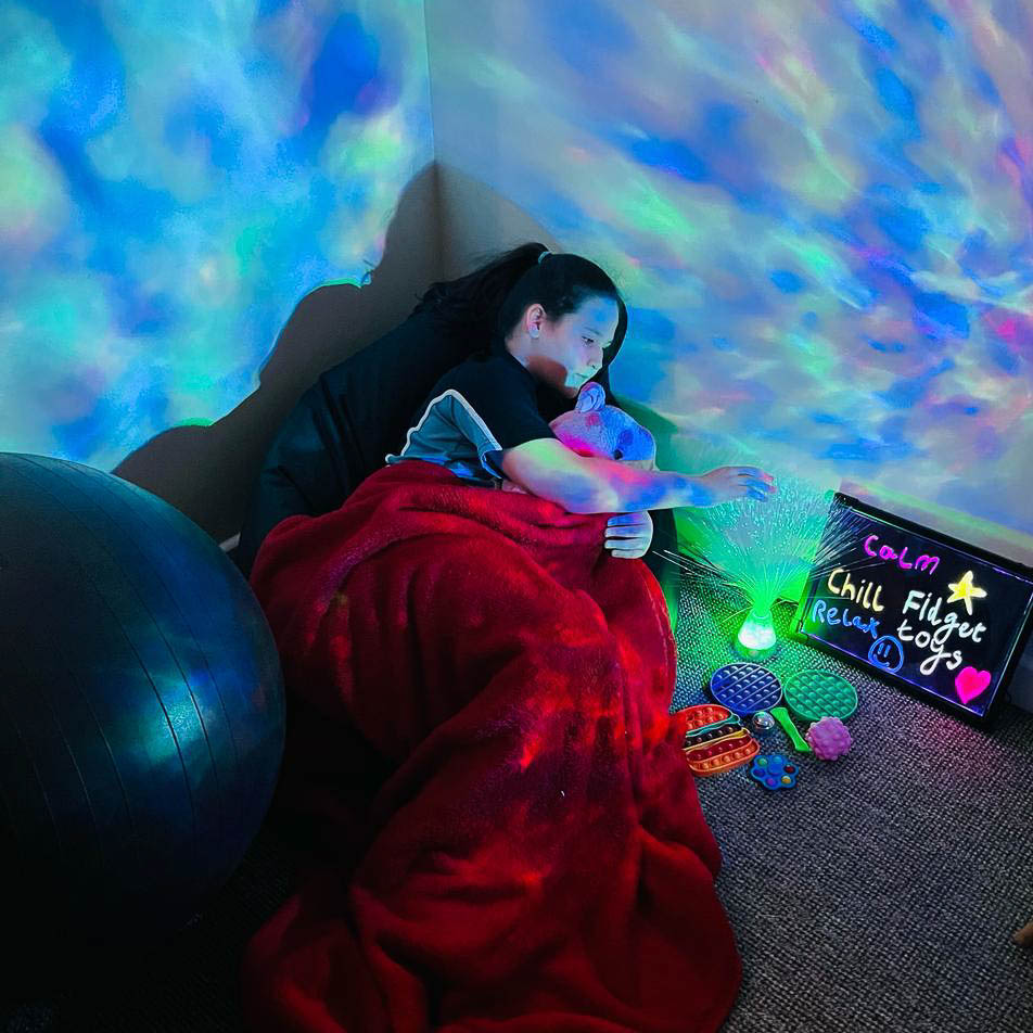 A child lays under a blanket in a sensory room. The lights are dim and they are touching an optical lamp and cuddling a soft toy.